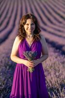 un mujer en un púrpura vestir es participación un ramo de flores de lavanda flores ella es sonriente y en pie en un campo de lavanda. foto