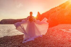 Mysterious woman silhouette long hair walks on the beach ocean water, sea nymph wind listens to the wave. Throws up a long white dress, a divine sunset. Artistic photo from the back without a face