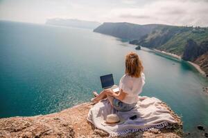 Lanza libre mujer trabajando en un ordenador portátil por el mar, mecanografía lejos en el teclado mientras disfrutando el hermosa vista, destacando el idea de remoto trabajar. foto