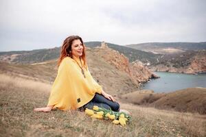 un mujer se sienta en el césped con un ramo de flores de amarillo flores en su regazo. ella es mirando fuera a el océano, tomando en el vista. concepto de paz y tranquilidad. foto