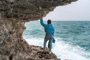 A woman in a blue jacket stands on a rock above a cliff above the sea and looks at the raging ocean. Girl traveler rests, thinks, dreams, enjoys nature. Peace and calm landscape, windy weather. photo