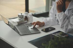 Serious female doctor using laptop and writing notes in medical journal sitting at desk. Young woman professional medic physician wearing white coat and stethoscope working on computer at workplace. photo