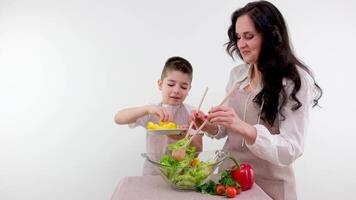 dentro a cozinha mãe e fofa pequeno Garoto cozinhando juntos saudável jantar. pais Ensinar pequeno filho saudável hábitos e quão para misturar legumes dentro a salada tigela. fofa criança ajudando dele pais video