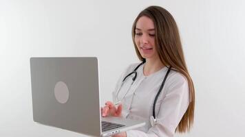 Portrait of beautiful senior doctor typing on laptop and turning to smile at camera. Close up of charming medical physician sitting in office and looking at camera video