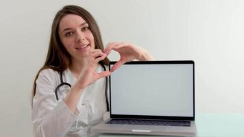 doctor shows a heart with her hands on the background of a white laptop monitor smile tenderness cleanliness and tidiness treatment care cardiology help people advertising new products medicines video
