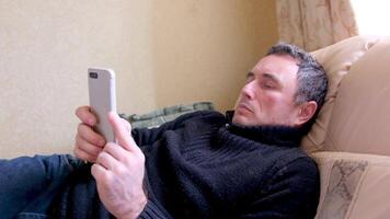 Closeup shot of a young man in mandatory quarantine. He stays in one room for two weeks. He lay on a bed and looks at the phone. He did not shave for a long time, he is stressed and upset. Staying in video