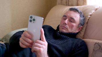 hombre utilizando iphone 13 Pro max en contra borroso un hombre mentiras en el cama descansando utilizando el teléfono teléfono inteligente en su manos cómodo relajado mentira descanso video