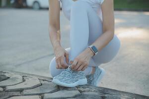 Mature fitness woman tie shoelaces on road. Cheerful runner sitting on floor on city streets with mobile and earphones wearing sport shoes. Active latin woman tying shoe lace before running. photo