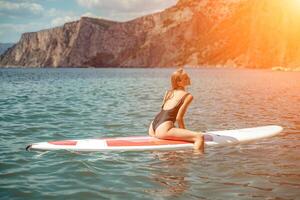 mujer cenar mar. Deportes niña en un tabla de surf en el mar en un soleado verano día. en un negro baños traje, él se sienta en un sapa en el mar. descanso en el mar. foto