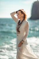 Woman beach sea white dress. The middle-aged looks good with blonde hair, boho style in a white long dress with beach decorations on the neck and arms. photo