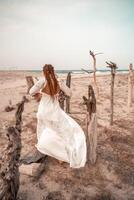 Model in boho style in a white long dress and silver jewelry on the beach. Her hair is braided, and there are many bracelets on her arms. photo