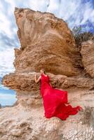 A woman in a red silk dress stands by the ocean, with mountains in the background, as her dress sways in the breeze. photo