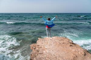 A woman in a blue jacket stands on a rock above a cliff above the sea and looks at the raging ocean. Girl traveler rests, thinks, dreams, enjoys nature. Peace and calm landscape, windy weather. photo