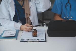 asiático psicólogo mujer señalando en pastillas botella a explicando medicina y prescripción a hembra paciente mientras dando asesoramiento acerca de médico y mental salud terapia a hembra paciente en clínica. foto