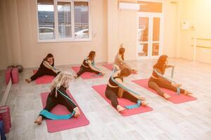 A group of six athletic women doing pilates or yoga on pink mats in front of a window in a beige loft studio interior. Teamwork, good mood and healthy lifestyle concept. photo