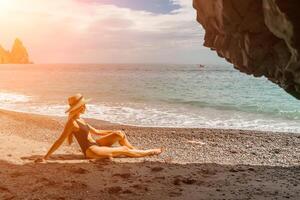 mujer viaje mar. ver de un mujer en un negro traje de baño desde un mar cueva atractivo mujer disfrutando el mar aire se sienta en el playa y mira a el mar. detrás su son rocas y el mar foto