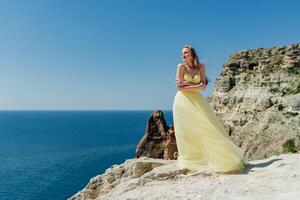 Woman in a yellow dress on the sea. Side view Young beautiful sensual woman in yellow long dress posing on a rock high above the sea at sunset. Girl in nature against the blue sky photo