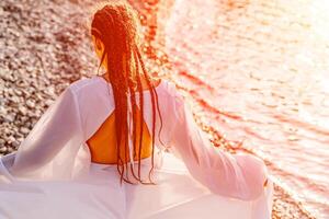 Mysterious woman silhouette long hair walks on the beach ocean water, sea nymph wind listens to the wave. Throws up a long white dress, a divine sunset. Artistic photo from the back without a face