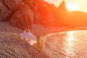 Mysterious woman silhouette long hair walks on the beach ocean water, sea nymph wind listens to the wave. Throws up a long white dress, a divine sunset. Artistic photo from the back without a face