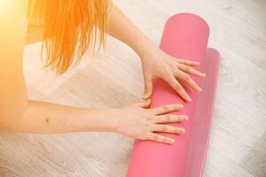 un joven mujer rollos un rosado aptitud o yoga estera antes de o después hacer ejercicio, hacer ejercicio a hogar en el vivo habitación o en un yoga estudio. sano hábitos, mantener adaptar, peso pérdida concepto. de cerca foto