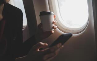 Blonde female tourist checking incoming notification on smartphone sitting on seat of airplane with netbook.Young businesswoman share media from telephone on laptop computer during plane flight photo