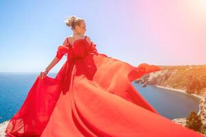 mujer mar rojo vestido. rubia con largo pelo en un soleado costa en un rojo fluido vestido, espalda vista, seda tela ondulación en el viento. en contra el fondo de el azul cielo y montañas en el costa. foto