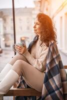 A middle-aged woman in a beige sweater with a blue mug in her hands is in a street cafe on the veranda photo