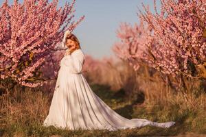 Woman blooming peach orchard. Against the backdrop of a picturesque peach orchard, a woman in a long white dress enjoys a peaceful walk in the park, surrounded by the beauty of nature. photo