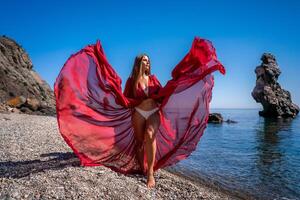 Beautiful sensual woman in a flying red dress and with long hair, stands on the seashore photo