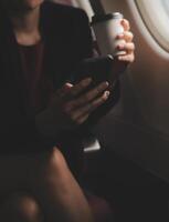Blonde female tourist checking incoming notification on smartphone sitting on seat of airplane with netbook.Young businesswoman share media from telephone on laptop computer during plane flight photo