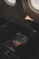 Blonde female tourist checking incoming notification on smartphone sitting on seat of airplane with netbook.Young businesswoman share media from telephone on laptop computer during plane flight photo