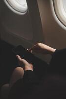 Blonde female tourist checking incoming notification on smartphone sitting on seat of airplane with netbook.Young businesswoman share media from telephone on laptop computer during plane flight photo