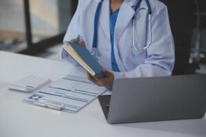 un profesional y enfocado asiático hembra médico en matorrales es trabajando y leyendo médico investigación en su ordenador portátil en su oficina a un hospital. foto