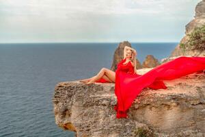 A woman in a red silk dress sits by the ocean with mountains in the background, her dress swaying in the wind. photo