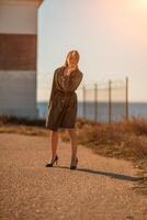 retrato rubia mar capa. un calma joven rubia en un caqui impermeable soportes en el costa en contra el fondo de un faro. foto