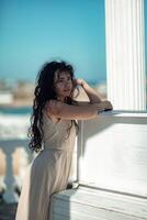 Sea woman rest. Portrait of a woman with long curly black hair in a beige dress stands on a balcony against the backdrop of the sea. Tourist trip to the sea. photo