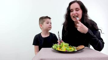 skön kvinna i svart skjorta och svart förkläde undervisning henne son på vilket sätt till ordna falsk frukt och grönsaker för dekoration i vit rena kök video