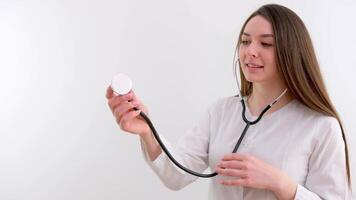 médical Université fille étudiant apprend à utilisation une phonendoscope correctement elle détient dans sa mains examine souriant dans une blanc manteau sur une blanc Contexte formation futur médecin être santé video