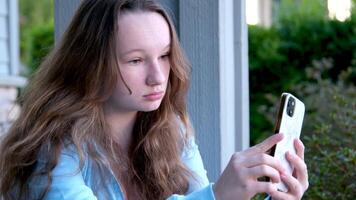 Closeup portrait child. Girl using mobile phone and smiling. Looking at camera. teenager girl straightens hair sits on doorstep of the house on the street admires herself in reflection of the mirror video