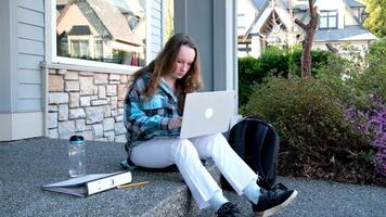a teenage girl is studying sitting against the background of gray house in laptop and typing work there is folder and water next to study online study online work first job school university exam video