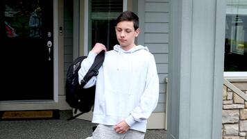 das Junge setzt ein Aktentasche auf seine Schultern ist vorbereiten zu gehen zu Schule gegen das Hintergrund von das Haus das Privat Sektor sieht aus um gekleidet im ein Sport Uniform Teenager video