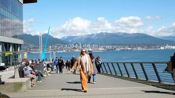 Canada Place a bright woman with a big scarf, a jacket, orange pants, sunglasses walks past the camera, couples sit on benches, Pacific Ocean mountains and statue of a drop in Vancouver video