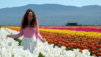 coleção do videos e fotos Onde uma menina dentro uma branco saia anda em através campo com tulipas azul fundo céu montanhas brilhante floração Campos adolescente jovem mulher andar ao ar livre beleza autocuidados