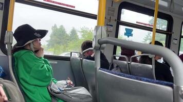 Extravagante mujer en un verde chaqueta y sombrero comiendo chocolate barras en un autobús sentado hablando en el teléfono solo en dos asientos con un bolso en su manos vida en el ciudad de Vancouver surrey Canadá 2023 video