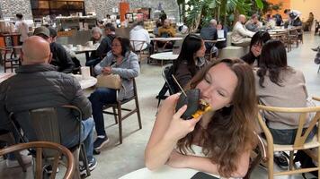 Adolescente niña joven mujer felizmente picaduras un negro hamburguesa bollo en un restaurante muchos personas delicioso comida sentar solo a mesa en real hora seco apagado lamer labios sonrisa gastar hora en un café Vancouver Canadá 2023 video