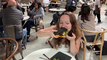 Tonårs flicka ung kvinna lyckligt biter en svart burger bulle i en restaurang många människor utsökt mat sitta ensam på tabell i verklig tid torr av slicka mun leende spendera tid i en Kafé vancouver kanada 2023 video