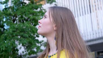 joven hermosa mujer, joven niña en grande ciudad en amarillo bordado camisa, bordado blusa, largo marrón cabello, sonrisa, pluma pendientes, amarillo y azul colores en ropa, ucranio nacional disfraz video
