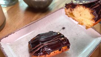 cut a piece of custard cake with butter cream poured over chocolate on a white plate in a restaurant, lift the piece with a knife and fork video