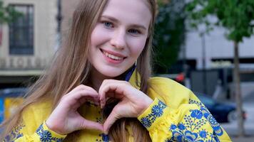 sorridente jovem menina dentro amarelo suéter mostrando coração com dois mãos, amor placa. amarelo Vyshyvanka ucraniano tradicional roupas dentro a grande cidade jovem mulher jovem amor para dela país Paz video