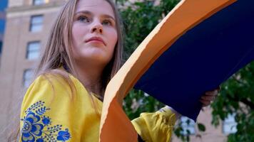 a beautiful Ukrainian woman in an embroidered holds Ukrainian flag on the background of the house a tree dreams of victory protection asks for help sad eyes fair hair confident strong imperious look video